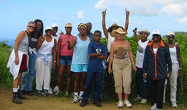 A group of people in hiking clothes stand for a group photo at a high vantage point overlooking Mount Victory Farm valley and the Caribbean sea.