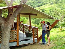Camp owner Bruce Wilson chats with a camper by the entrance to the deck of the teak bungalow at Mt. Victory Camp, with the view of the green farm valley in the distance.